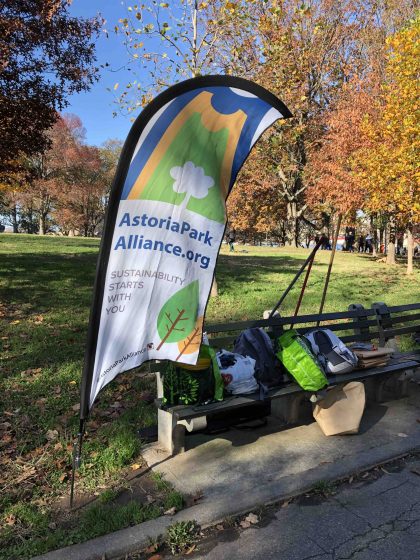Banner that reads "AstoriaParkAlliance.org Sustainability Starts with You". The banner is near a bench with a variety of backpack and bags.